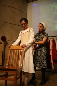 Lindsay has slipped on the corset and waits for Kitty to lace her up.